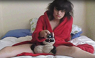 Woman lying in bed, wearing a red robe, cradling a stuffed cat and holding a remote control